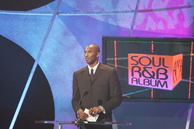 A bald Black man in a black three-button suit holds an envelope while at an on-stage podium, with "Soul R&B Album" projected on a screen behind him.