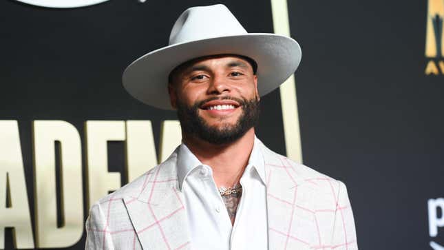 A Black man in a white broad-brimmed hat and pink and white suit coat with pink plaid smiles at the camera.